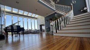 Ein geräumiges Wohnzimmer mit einem Flügel, einer Treppe mit weißen Balustraden und großen Fenstern, die viel natürliches Licht hereinlassen.