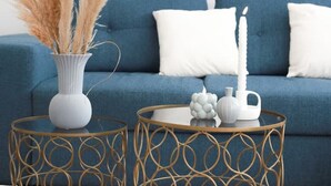 A blue sofa with white cushions, a white vase with pampas grass, a white candle, and two round tables with gold wire designs. - L'amiral Centre Ville Balnéo Parking (Grasse)