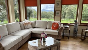 A modern living room with a sectional sofa, a glass coffee table, and a view of a lush green landscape through large windows. - SECLUDED COTTAGE IN THE VERY HEART OF THE HIGHLANDS OF SCOTLAND (Kingussie)