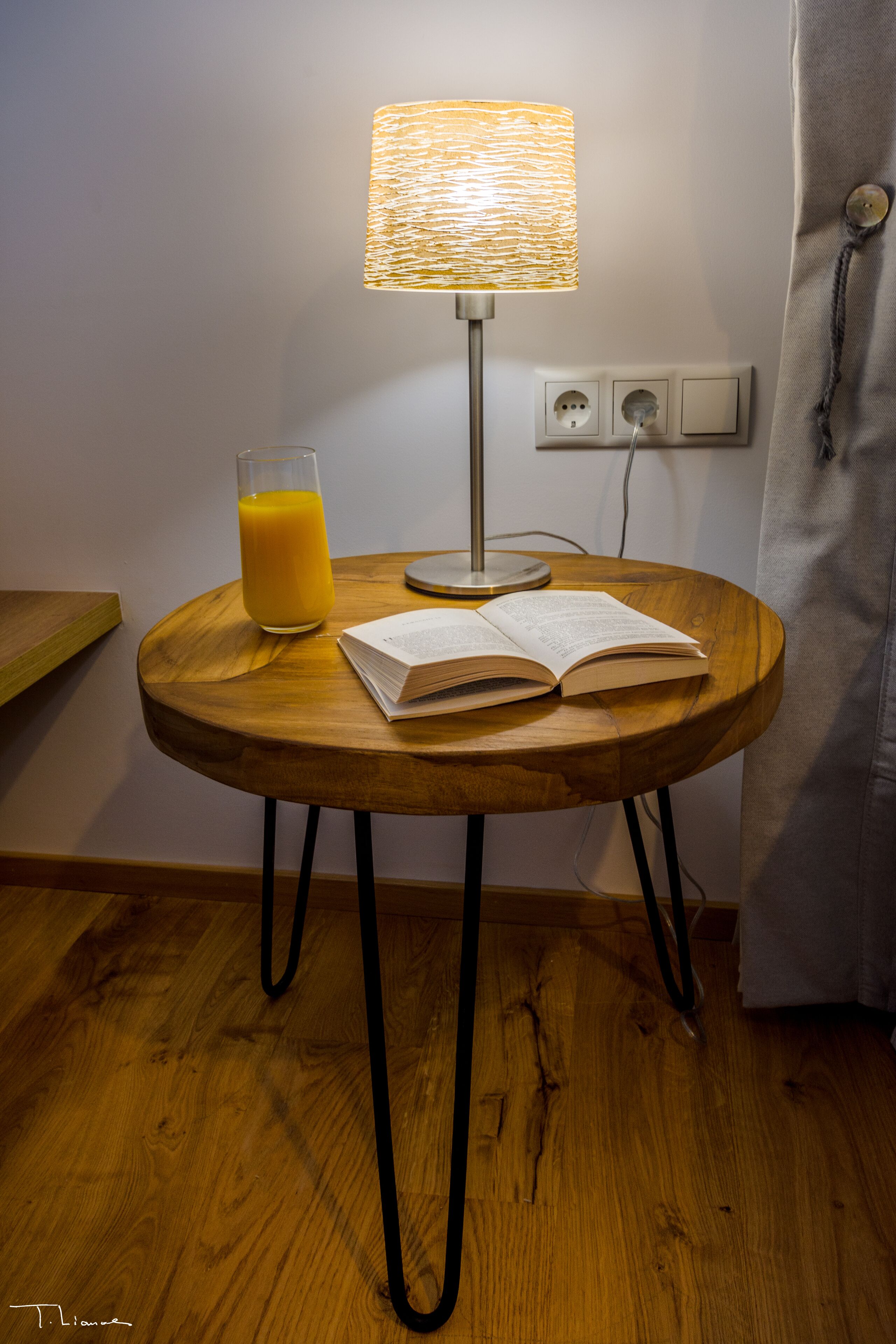 A round wooden table with a lamp, a glass of orange juice, and an open book.