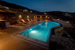 A swimming pool with lounge chairs and a stone wall, surrounded by a hillside.