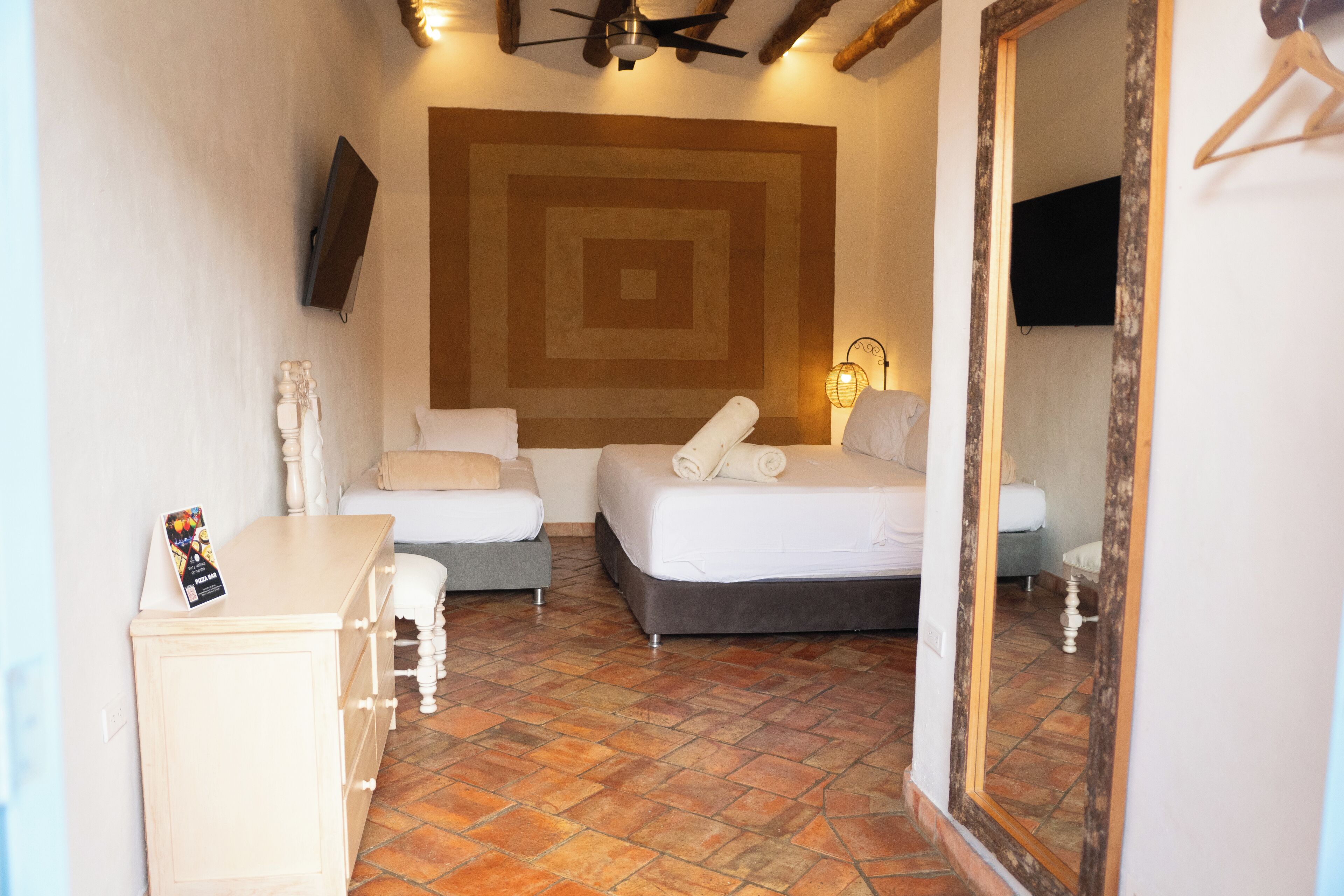 Habitación de hotel con una cama grande, una cama más pequeña, un escritorio y un aparador. Hay un espejo grande en la pared y un televisor montado sobre la cama.