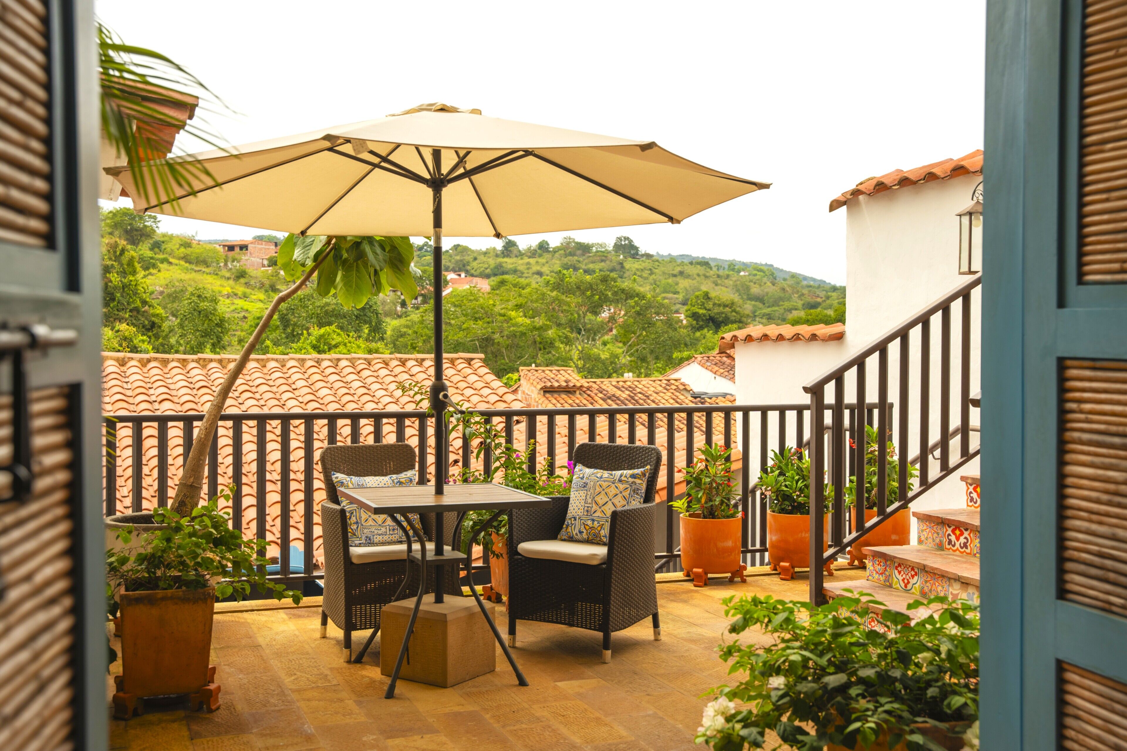 Um pátio externo com mesa e cadeiras, cercado por plantas em vasos e um grande guarda-sol.