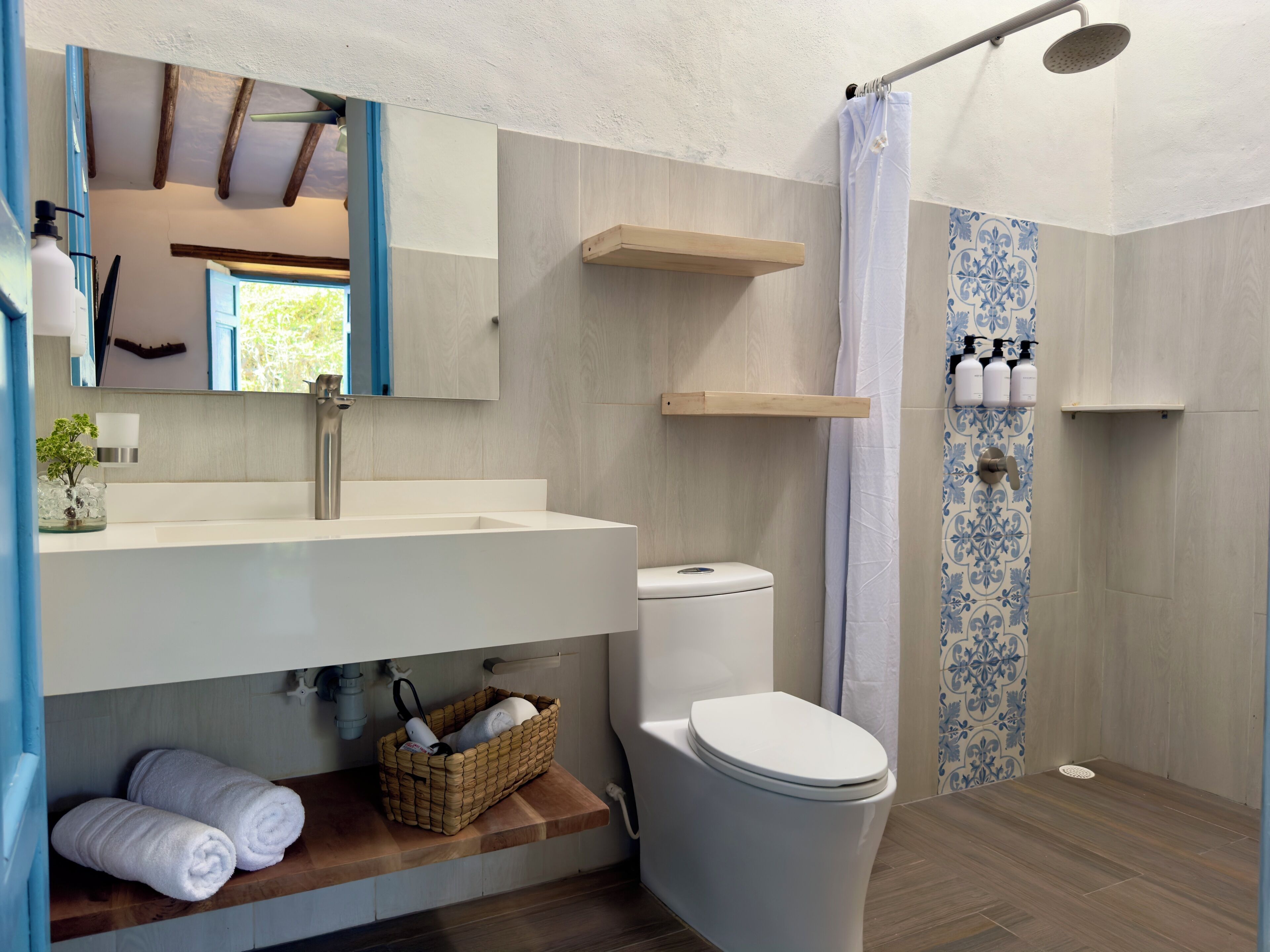 A modern bathroom with a large sink, toilet, and shower area. There are shelves above the sink and a basket with towels on the floor.