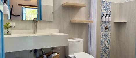 A modern bathroom with a large sink, toilet, and shower area. There are shelves above the sink and a basket with towels on the floor.