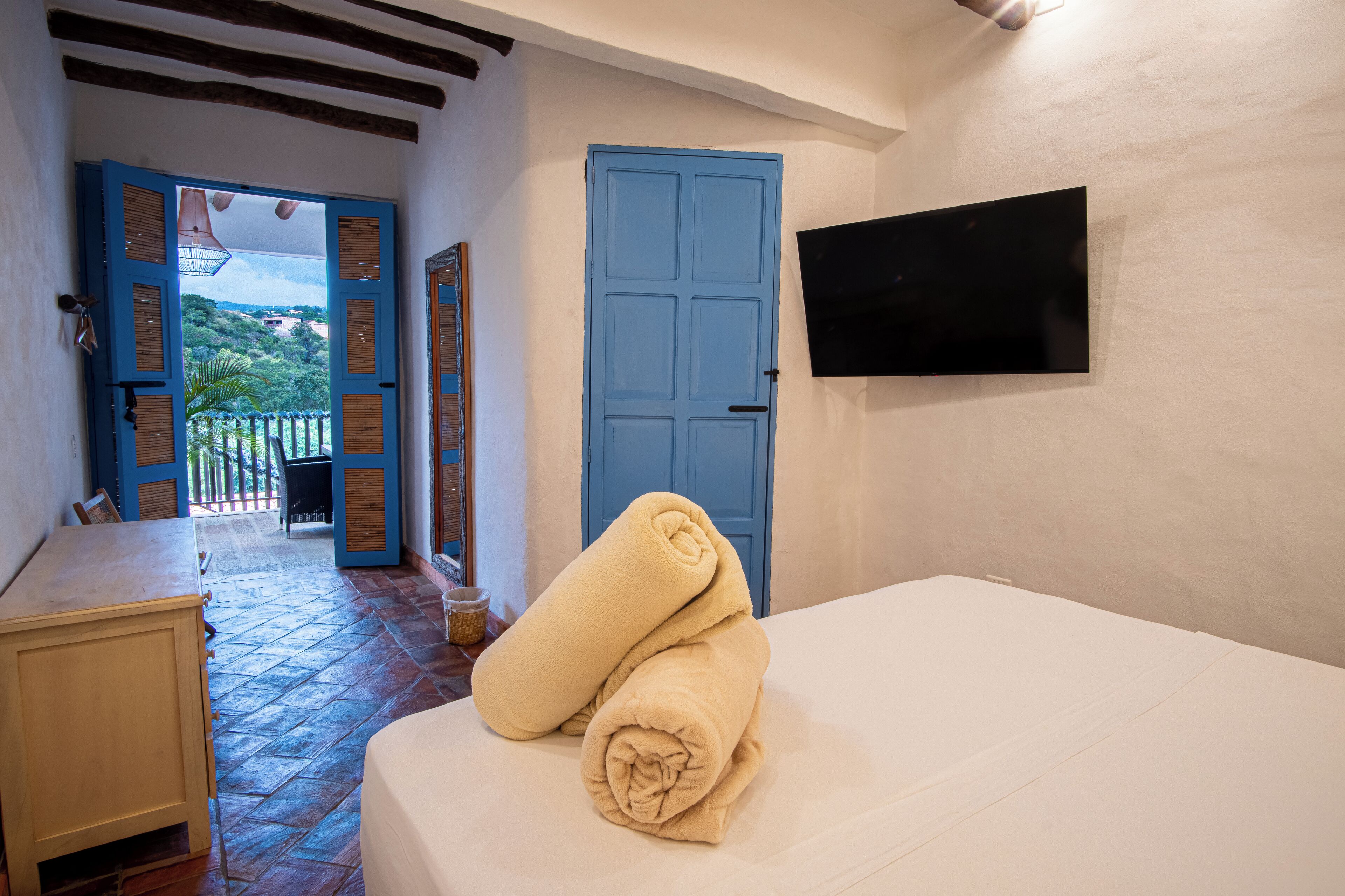 A room with a bed, a blue door, a TV mounted on the wall, and a view of a balcony with a railing.
