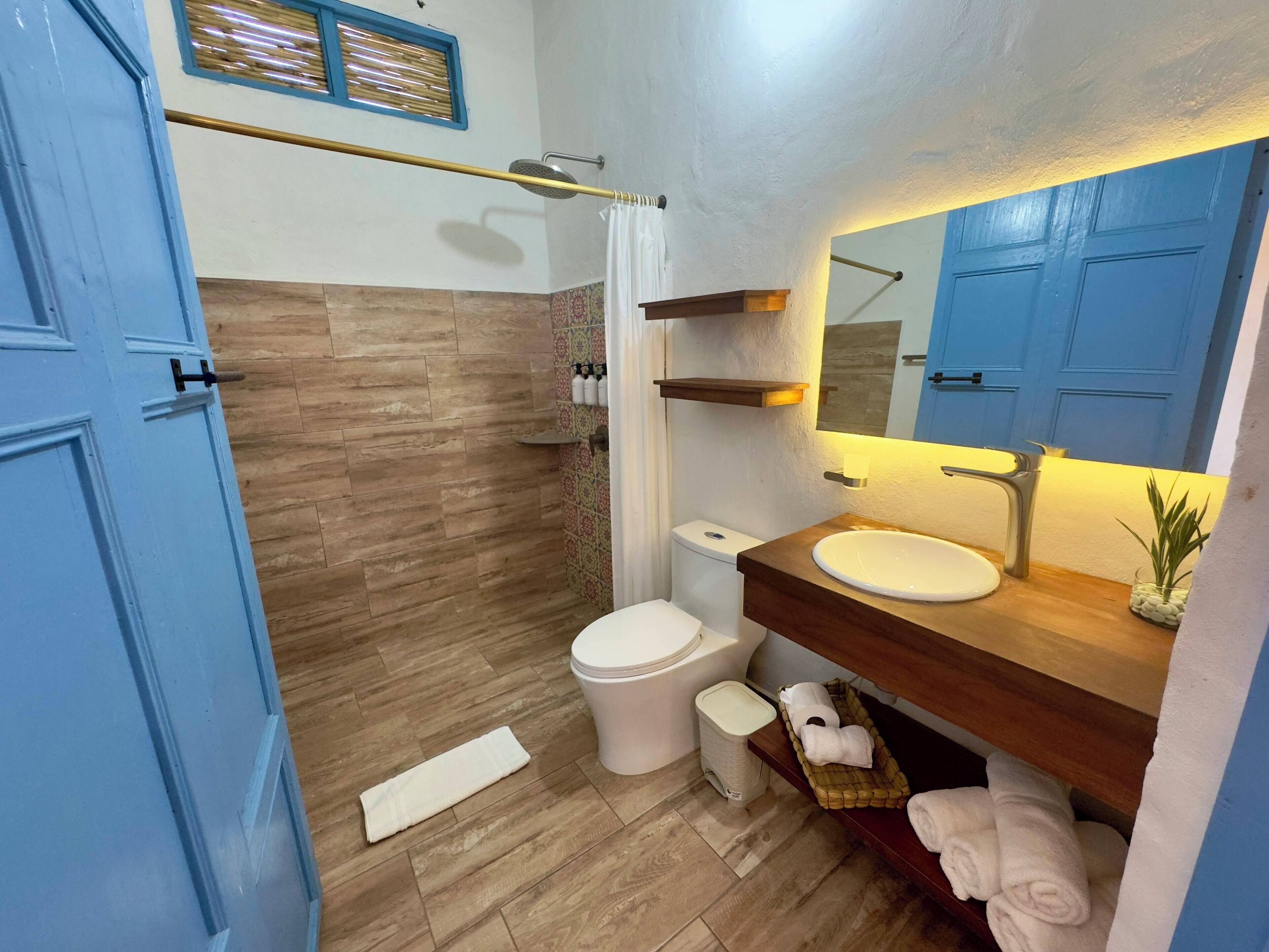 A modern bathroom with a wooden floor, a white toilet, a sink with a wooden countertop, a shower with a glass door, and a blue door.