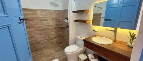 A modern bathroom with a wooden floor, a white toilet, a sink with a wooden countertop, a shower with a glass door, and a blue door.