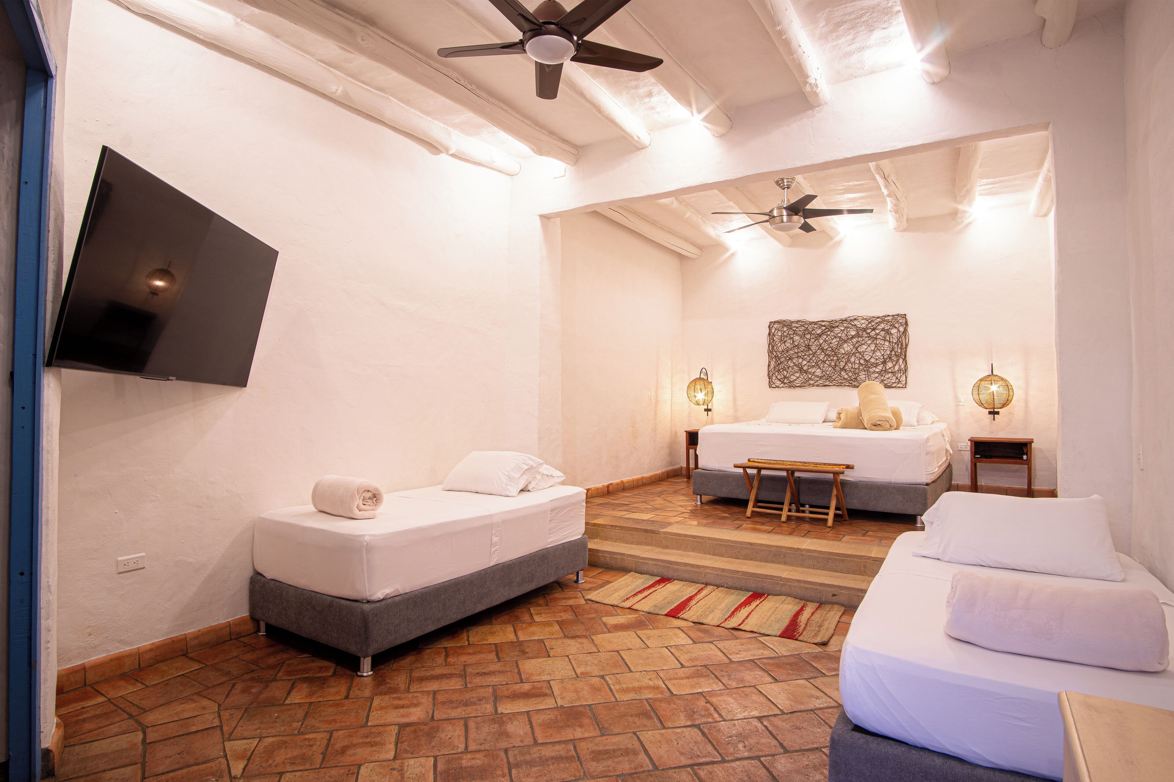 Habitación de hotel con dos camas, un televisor de pantalla plana montado en la pared, un ventilador de techo y un tapiz mural estampado.