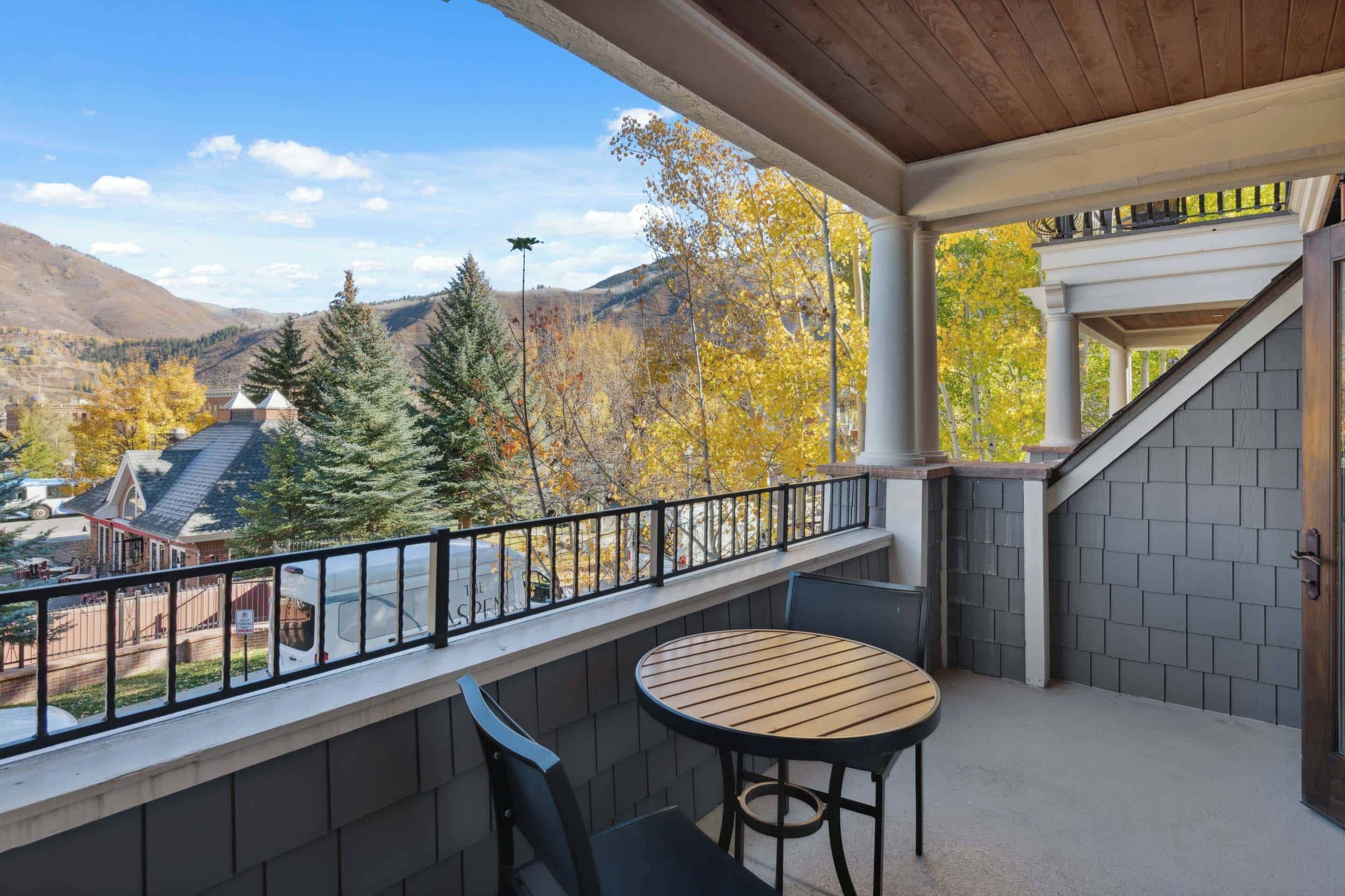 A balcony with a table and chairs, overlooking a mountainous landscape with autumn foliage.