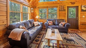 A log cabin interior with two leather sofas, a wooden coffee table, and large windows offering forest views. - Hot tub, Firepit, Beautiful Mountain Views! (Stanardsville)