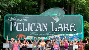 Un grupo de personas frente a un cartel de bienvenida a Lago Pelican, con un gráfico de un pez grande y árboles de fondo.