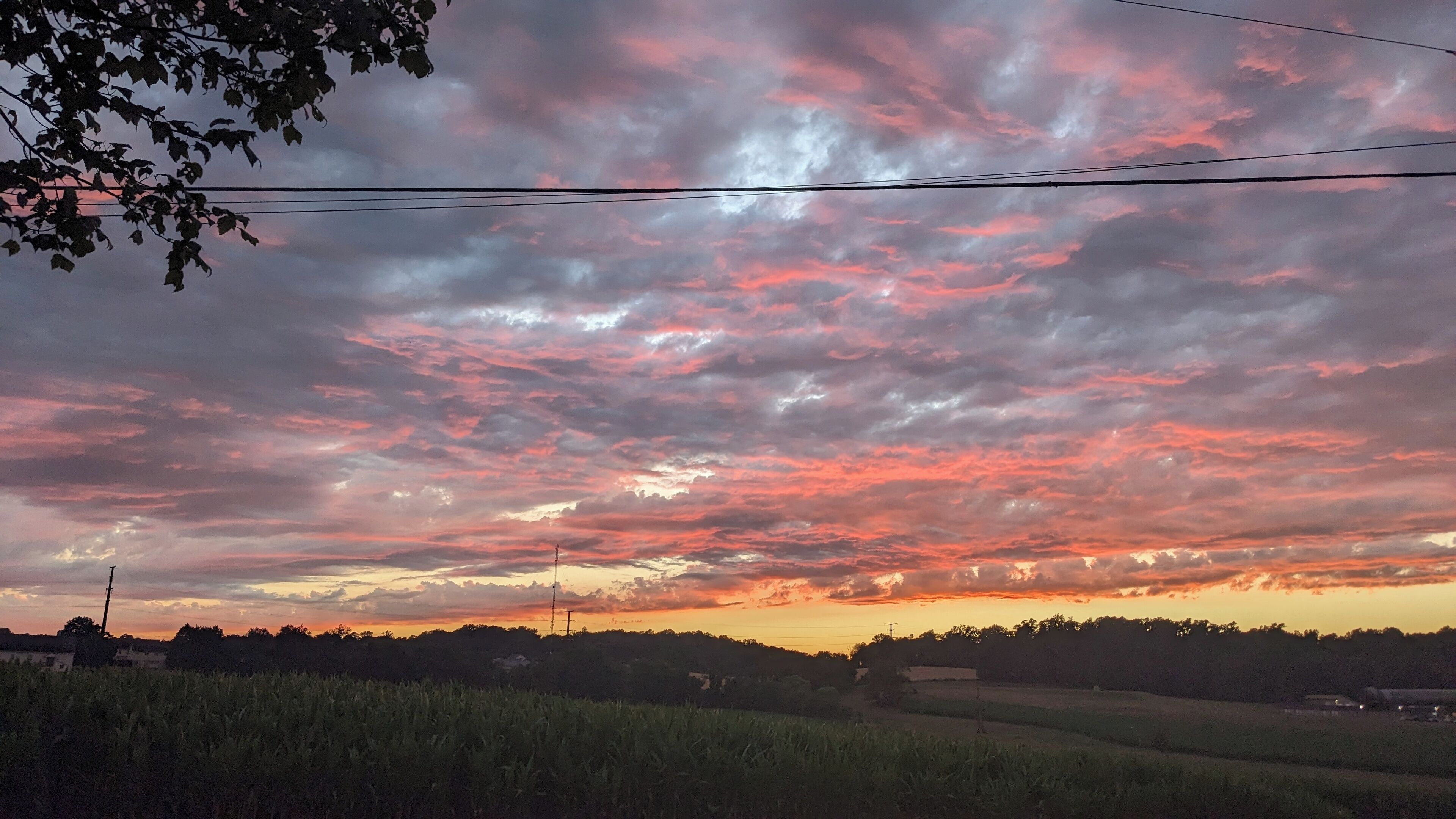 Auringonlasku maaseutumaisemassa, jossa on peltoja ja sähkölinjoja.