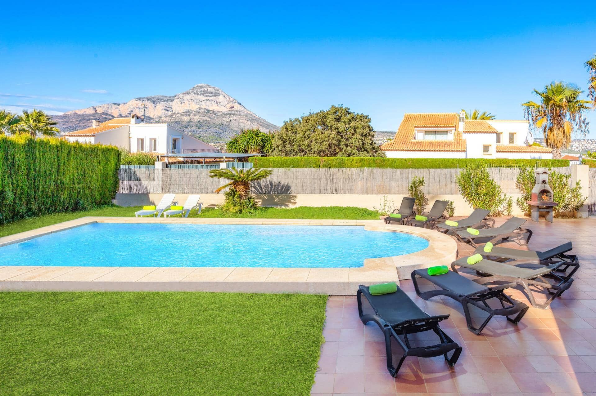 A swimming pool with lounge chairs and a barbecue area, surrounded by a well-maintained lawn and a mountain view.