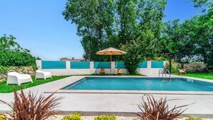 Une piscine bordée d’un muret en pierre, deux chaises longues blanches et un parasol.