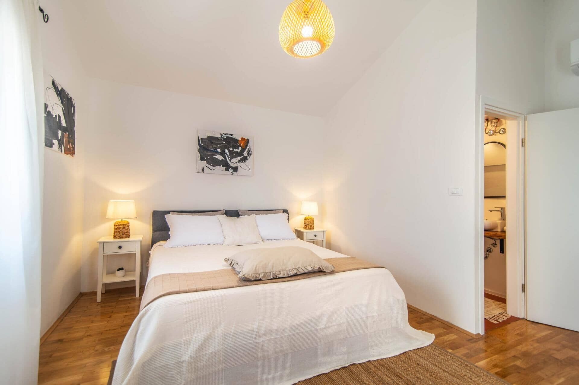 A bedroom with a bed, two bedside tables with lamps, a hanging light fixture, and a door leading to a bathroom.