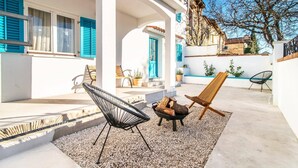 Une terrasse avec des sièges extérieurs, un foyer et un bâtiment blanc avec des volets bleus.