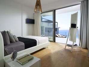 A modern hotel room with a bed, a bedside table with a book, a TV, and a balcony with a pool and lounge chairs.