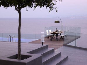 A modern outdoor dining area with a glass railing, a table set for two, and a view of the sea.