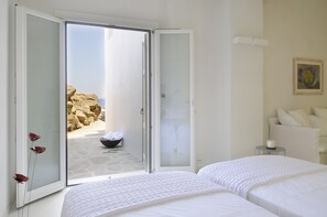 A hotel room with a bed, a bedside table with a candle, and a view of a rocky beach through an open door. - Inviting 7-bed Villa in Mikonos - Villa Jackie (Mykonos)
