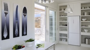 Une cuisine moderne avec un réfrigérateur blanc, des étagères ouvertes et une vue sur un espace extérieur aménagé avec des meubles blancs.