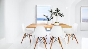 Un coin repas minimaliste avec une table en bois, des chaises blanches et une vue sur la mer grâce à une grande fenêtre.