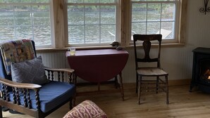 Una camera accogliente con pavimento in legno, una poltrona a dondolo, un tavolo rosso e vista su un fiume attraverso ampie finestre.