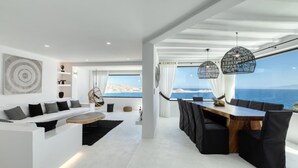 A modern living room with a large window overlooking the sea, a dining area with a wooden table and chairs, and a hanging chair. - Captivating 5-bed Villa in Mikonos Giullietta (Mykonos)