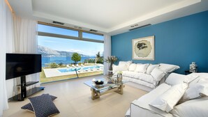 A spacious living room with a large window overlooking a pool and mountains. There is a white sofa, a glass coffee table, and a flat-screen TV.