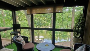 Una habitación con un ventanal grande, dos sillas de mimbre, una mesita redonda y vistas a un lago y árboles.