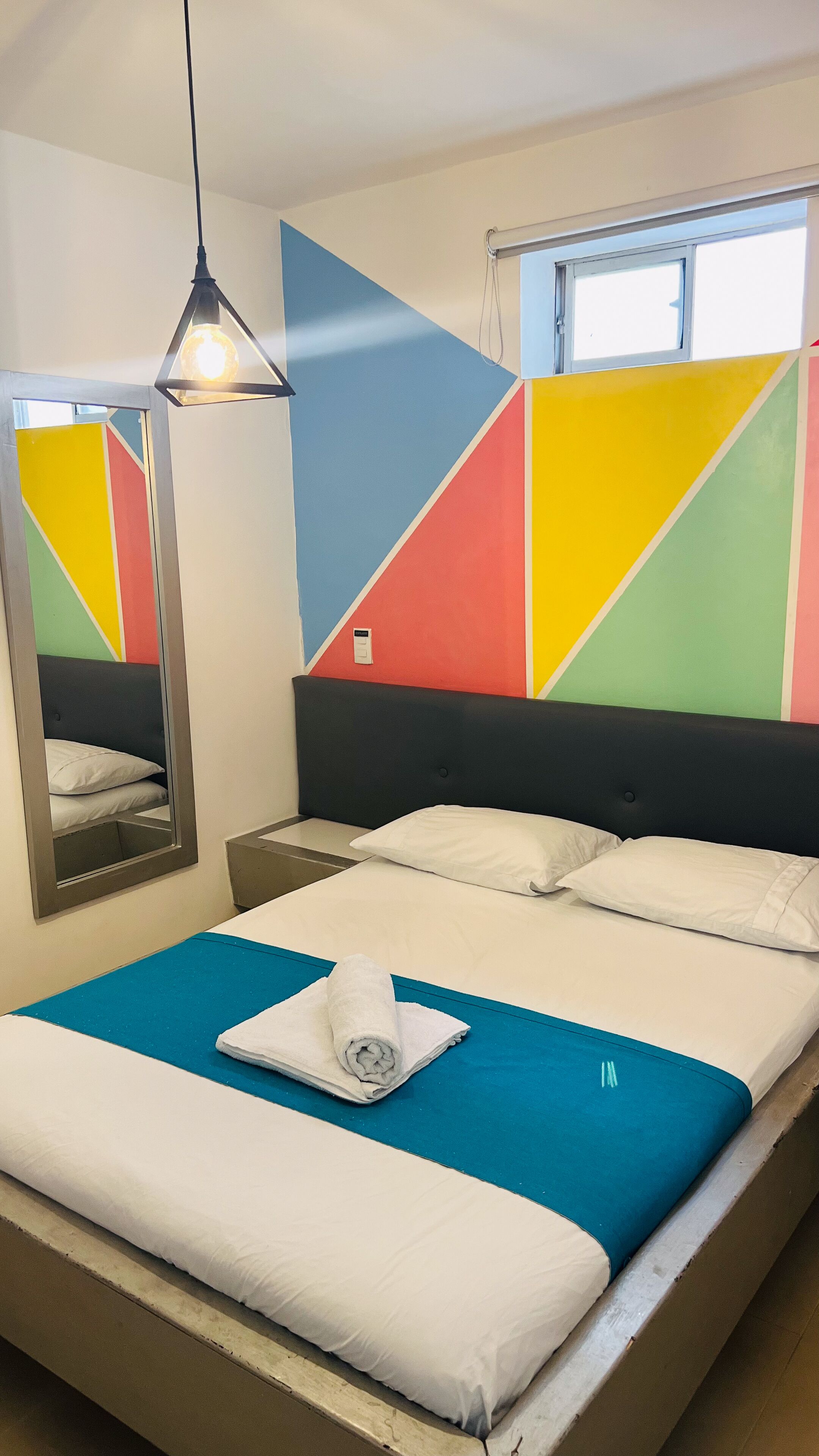 Une chambre d’hôtel avec un lit, une table de chevet, un miroir et un motif géométrique coloré sur le mur.