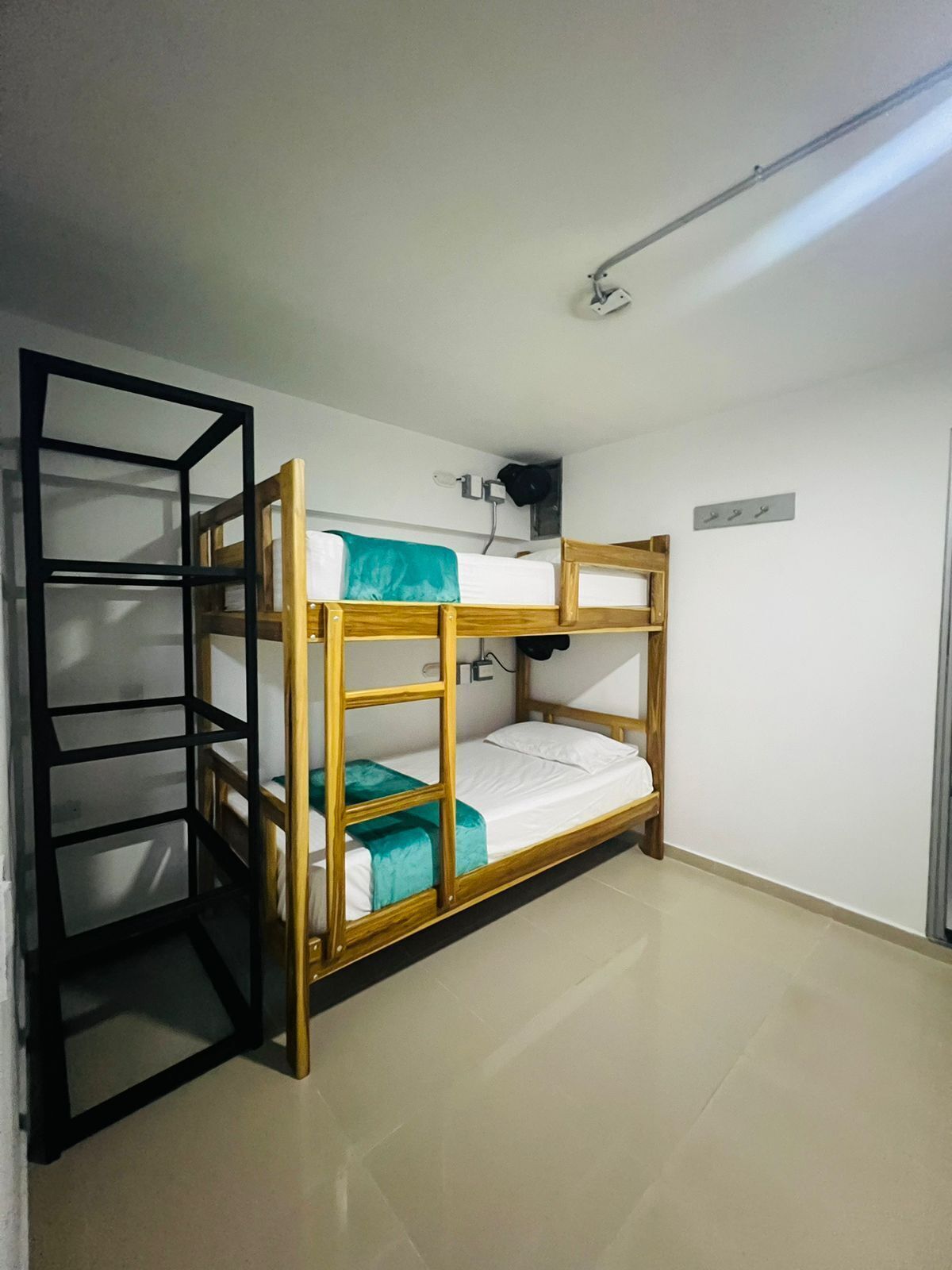 Ein Stockbett mit Holzrahmen und einer schwarzen Metallleiter. Eine weiße Wand mit einem kleinen Regal und einem Haken.