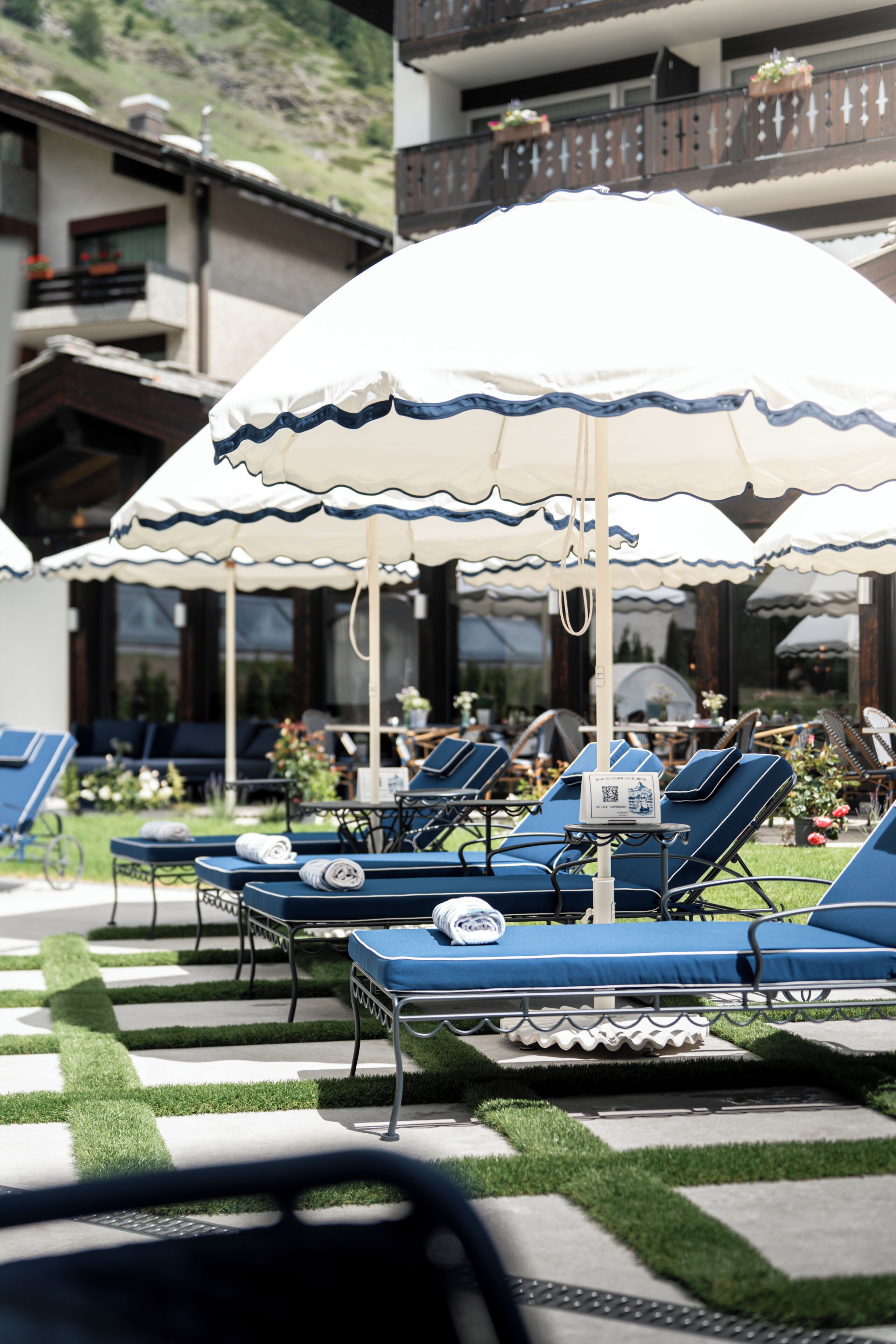 A poolside area with lounge chairs and umbrellas, a checkered pattern on the ground, and a building with balconies in the background.