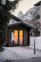 Uma cabana de madeira com porta de vidro, um roupão branco pendurado na porta e uma paisagem coberta de neve com montanhas ao fundo.