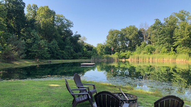 A serene lakeside setting with three Adirondack chairs and a small fire pit, surrounded by lush greenery and a calm pond. - Bloomington hideaway, 10 min from downtown, catch some fish in private pond. (Bloomington)