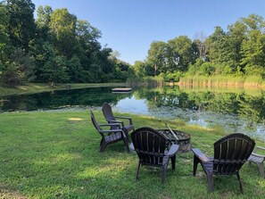Un cadre paisible au bord du lac, avec trois chaises Adirondack et un petit foyer, le tout entouré d’une végétation luxuriante et d’un  étang tranquille.