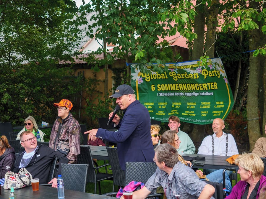 A group of people seated at an outdoor table with a banner in the background advertising a garden party.