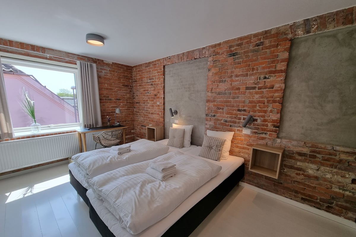 A modern bedroom with a brick wall, a large bed with white linens, a desk with a lamp, and a view of the outside.