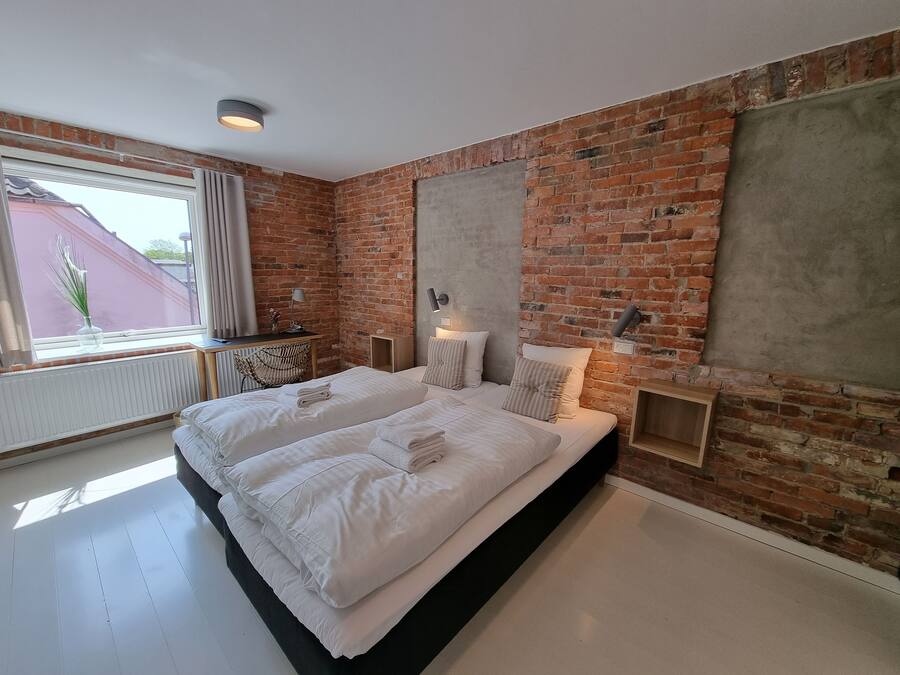 Un dormitorio moderno con pared de ladrillo, cama grande con ropa blanca, escritorio con lámpara y vista al exterior.
