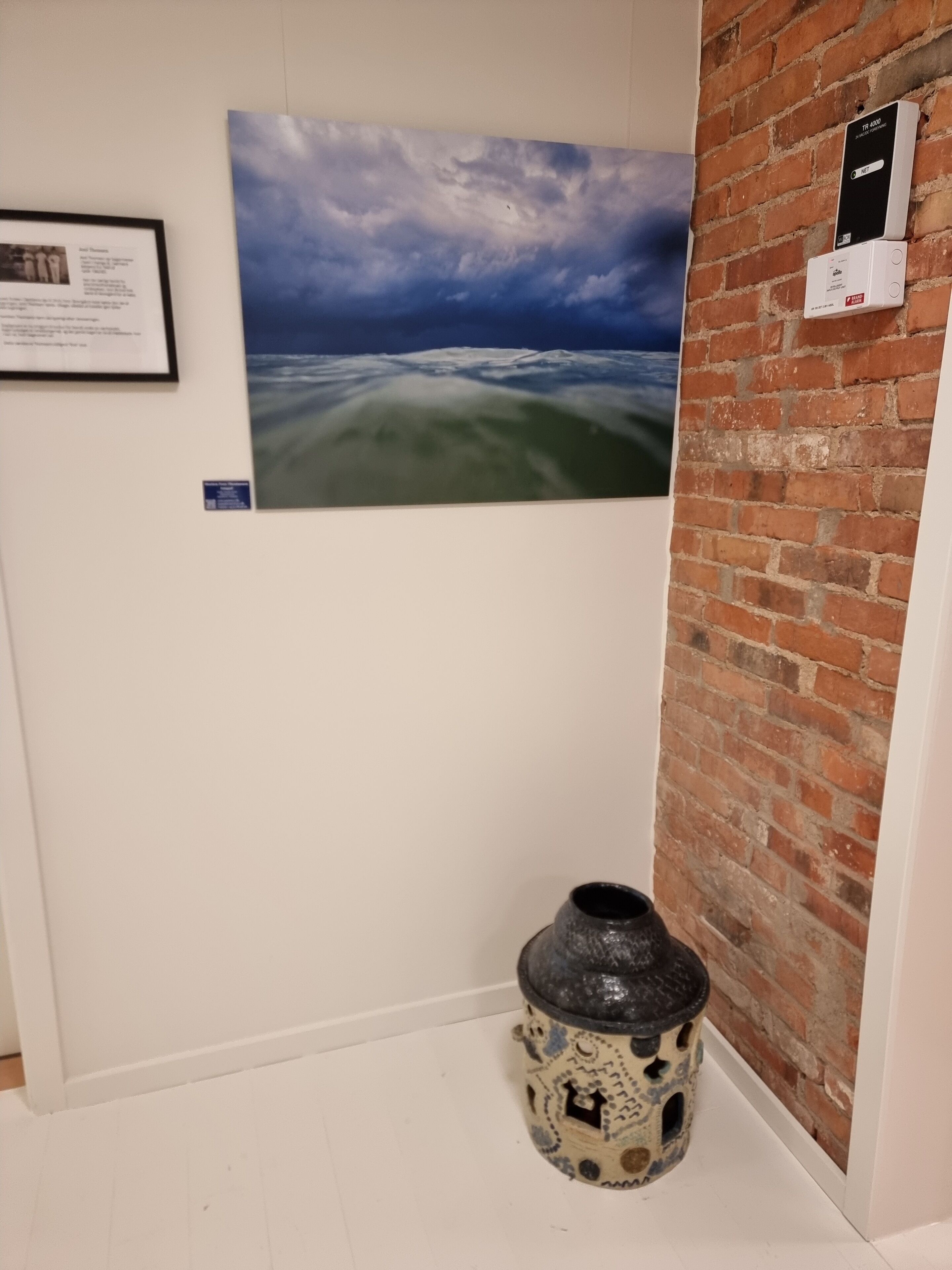A room with a brick wall, a framed picture of a landscape, a small decorative vase, and a thermostat on the wall.