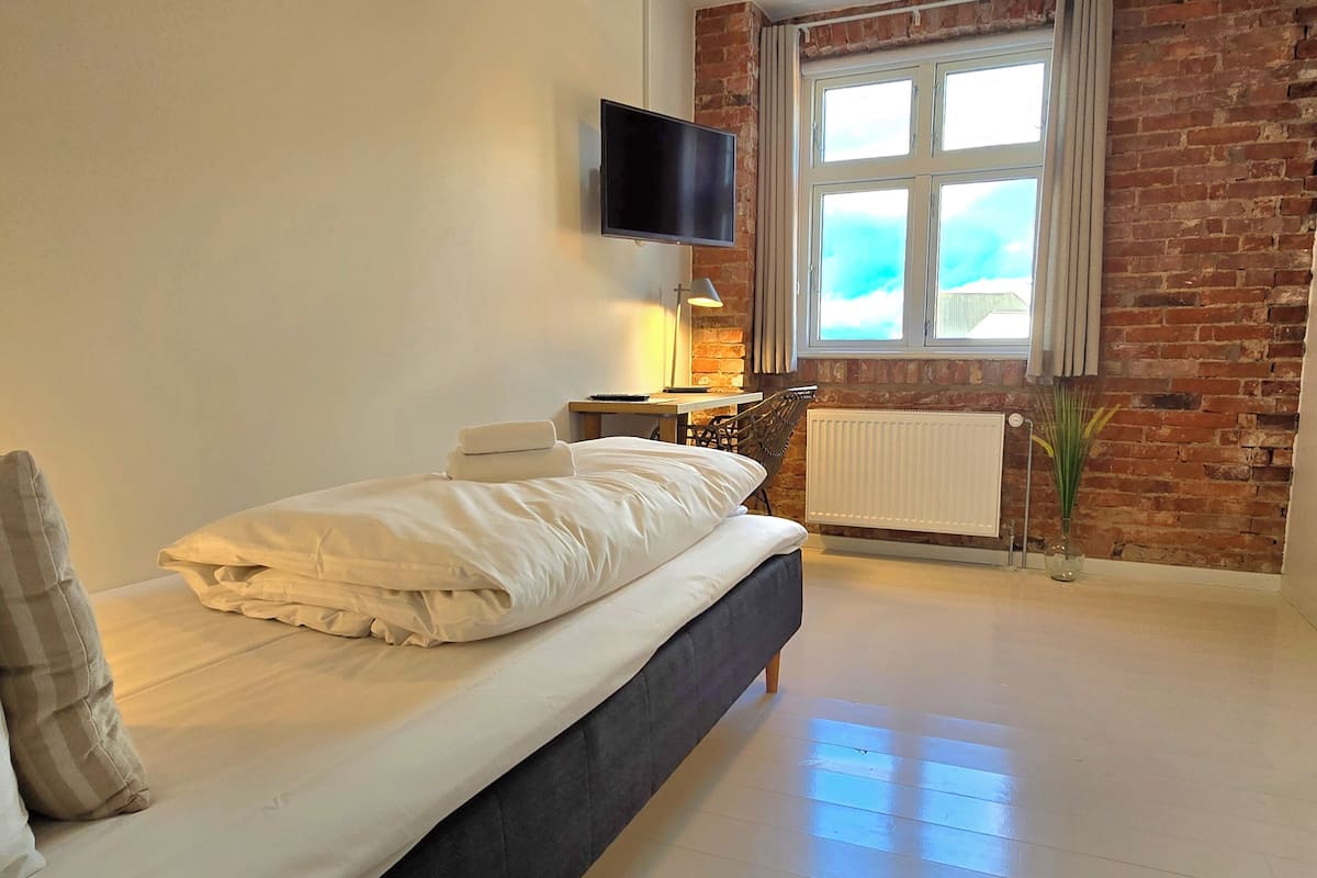 A modern bedroom with a bed, a TV mounted on the wall, a window with curtains, and a brick wall.