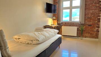 Un dormitorio moderno con cama, televisor montado en la pared, ventana con cortinas y pared de ladrillo.