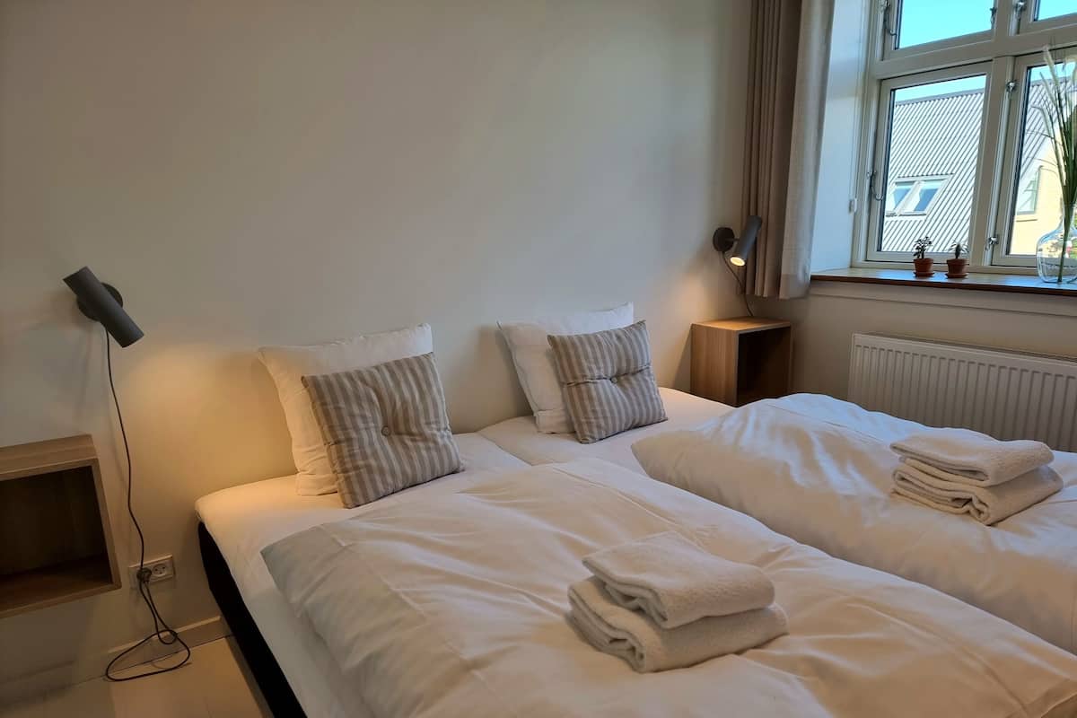 A neatly made double bed with white linens and two pillows, a bedside table with a lamp, and a window with curtains.