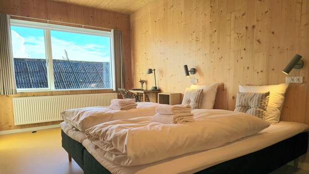 Una cama grande con sábanas blancas, cabecera de madera y vista a un edificio con tejado azul.