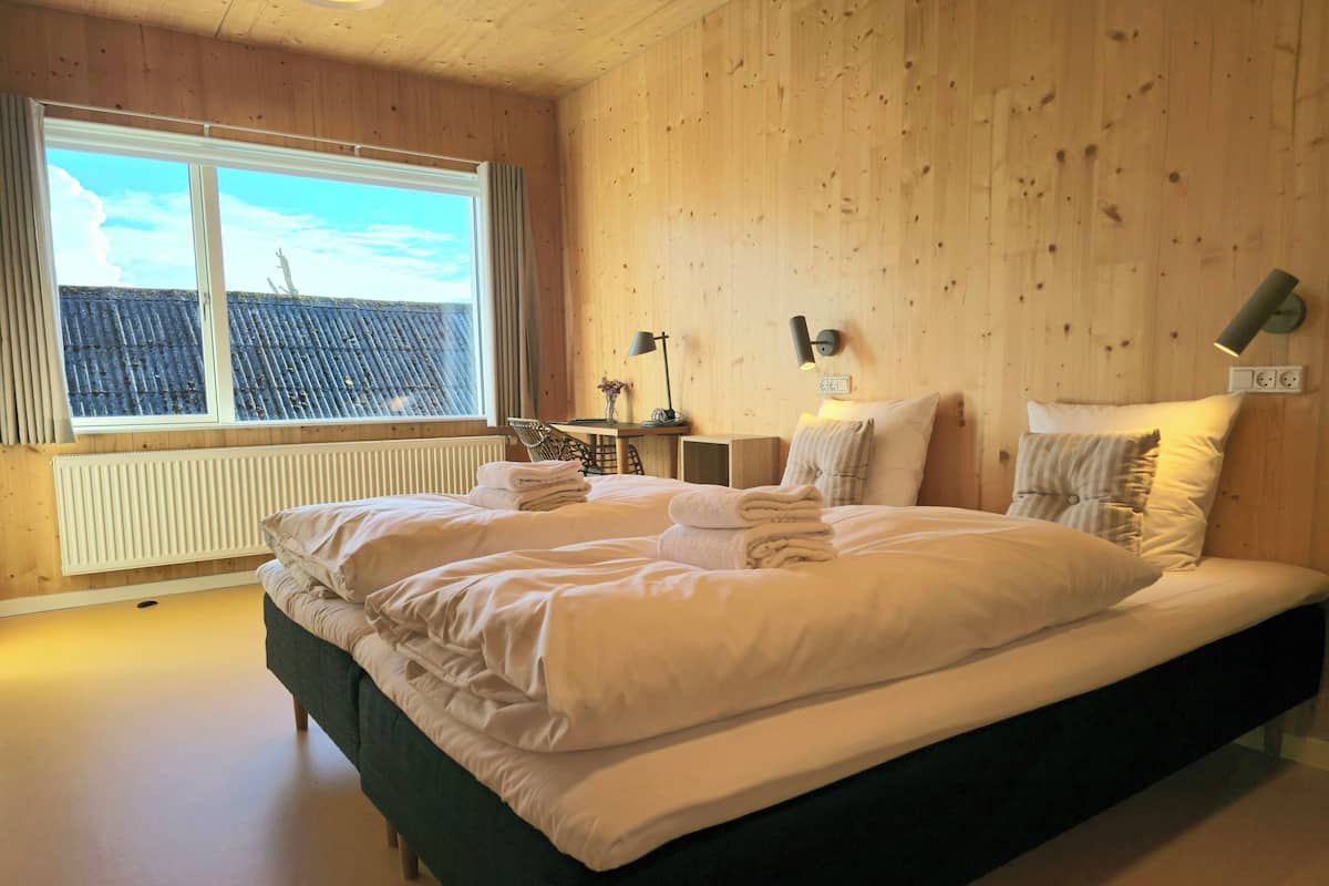 A large bed with white linens, a wooden headboard, and a view of a blue-roofed building outside the window.