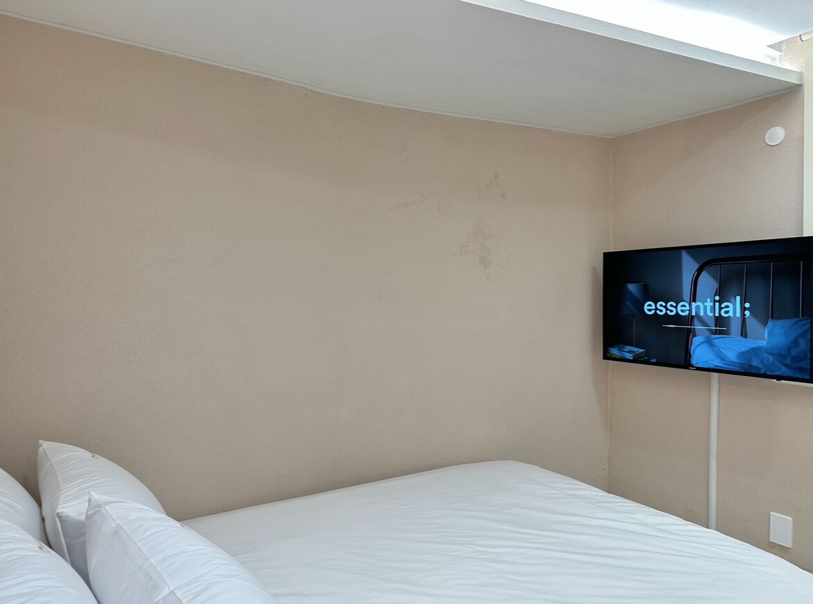 A hotel room with a bed, a television mounted on the wall, and a plain beige wall.