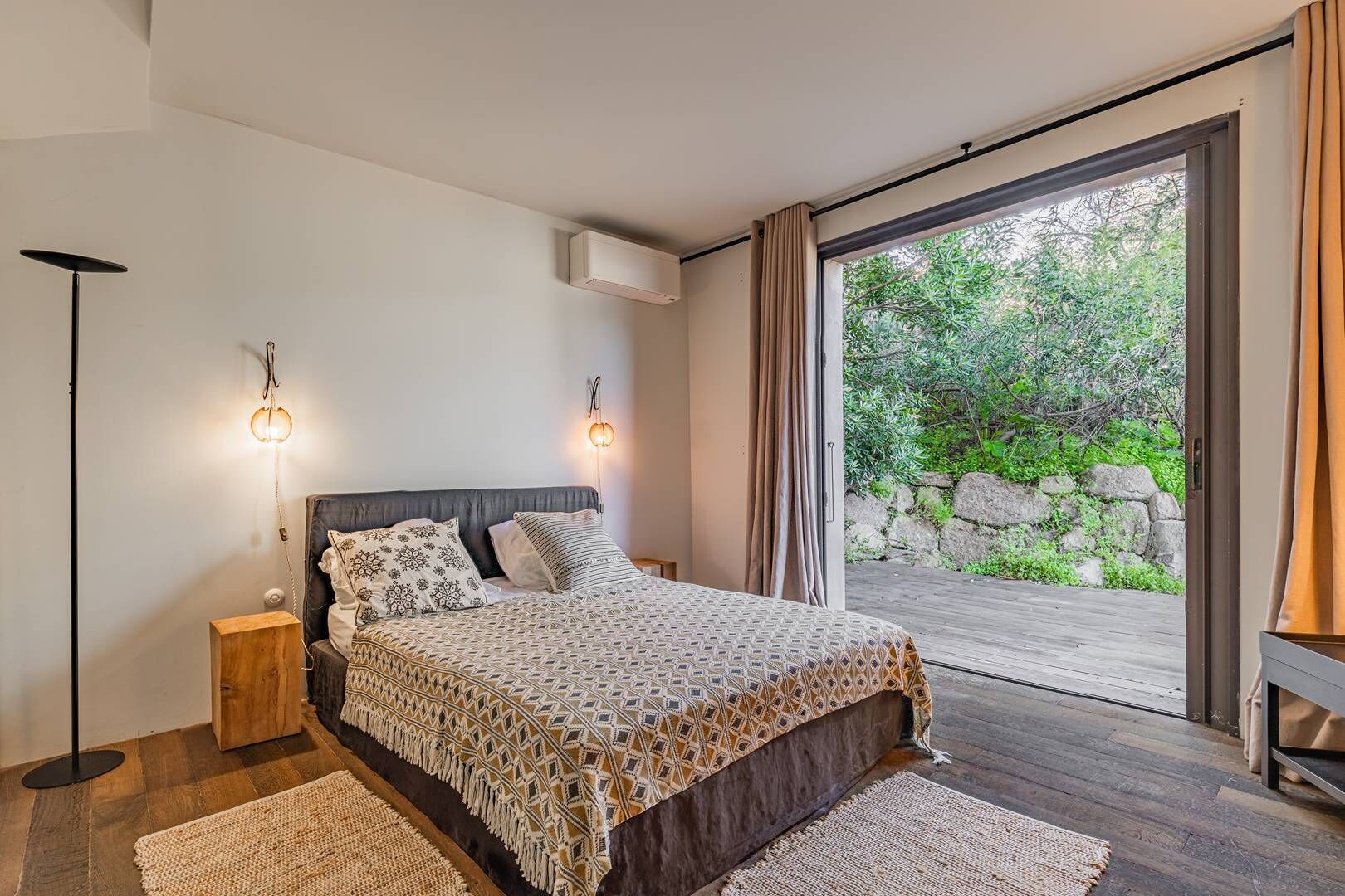 Une chambre avec un grand lit, deux lampes de chevet, un plancher en bois et une vue sur la verdure grâce à une porte coulissante.