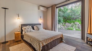 A bedroom with a large bed, two bedside lamps, a wooden floor, and a view of greenery through a sliding door.