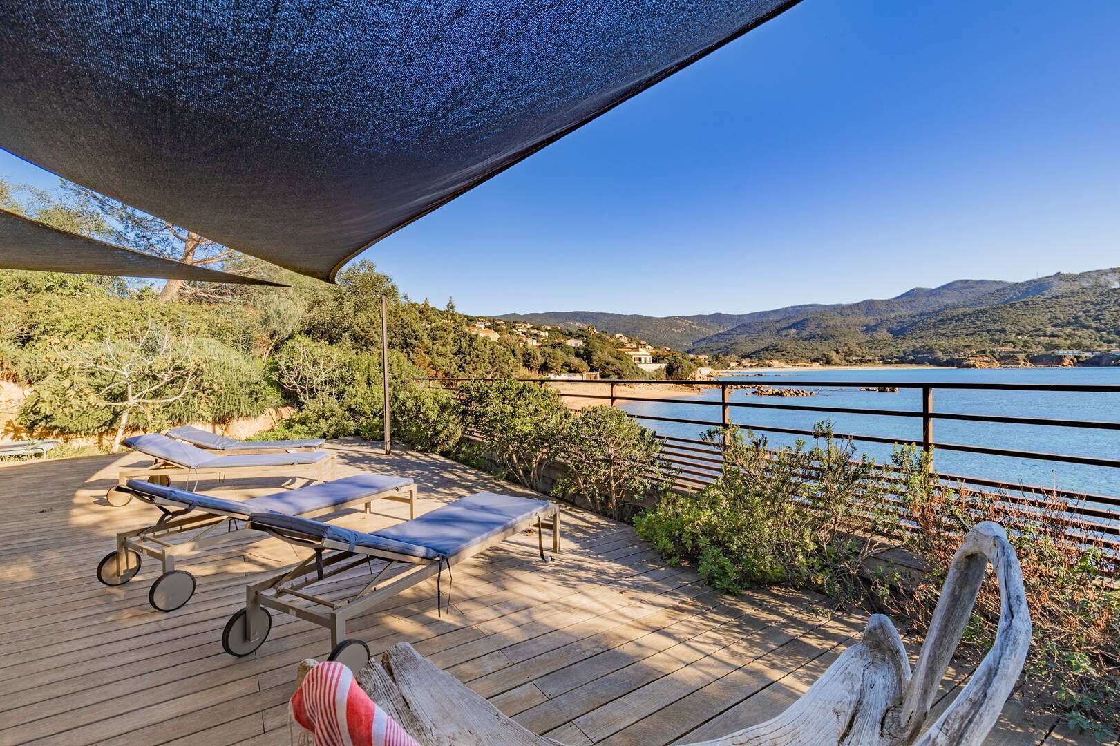 A wooden deck with sun loungers and a shaded area overlooking a body of water and hills.