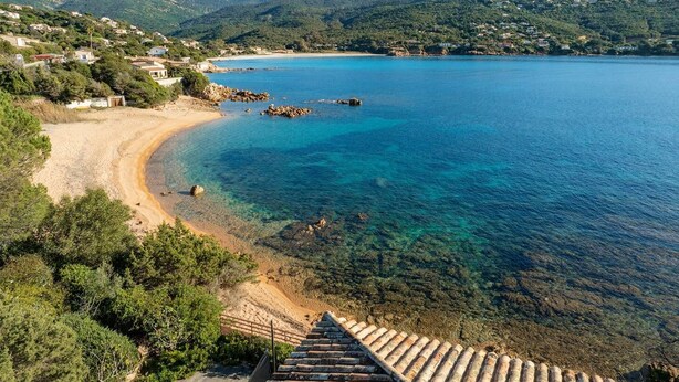 A coastal landscape with a clear beach, rocky outcrops, and a hilly backdrop.